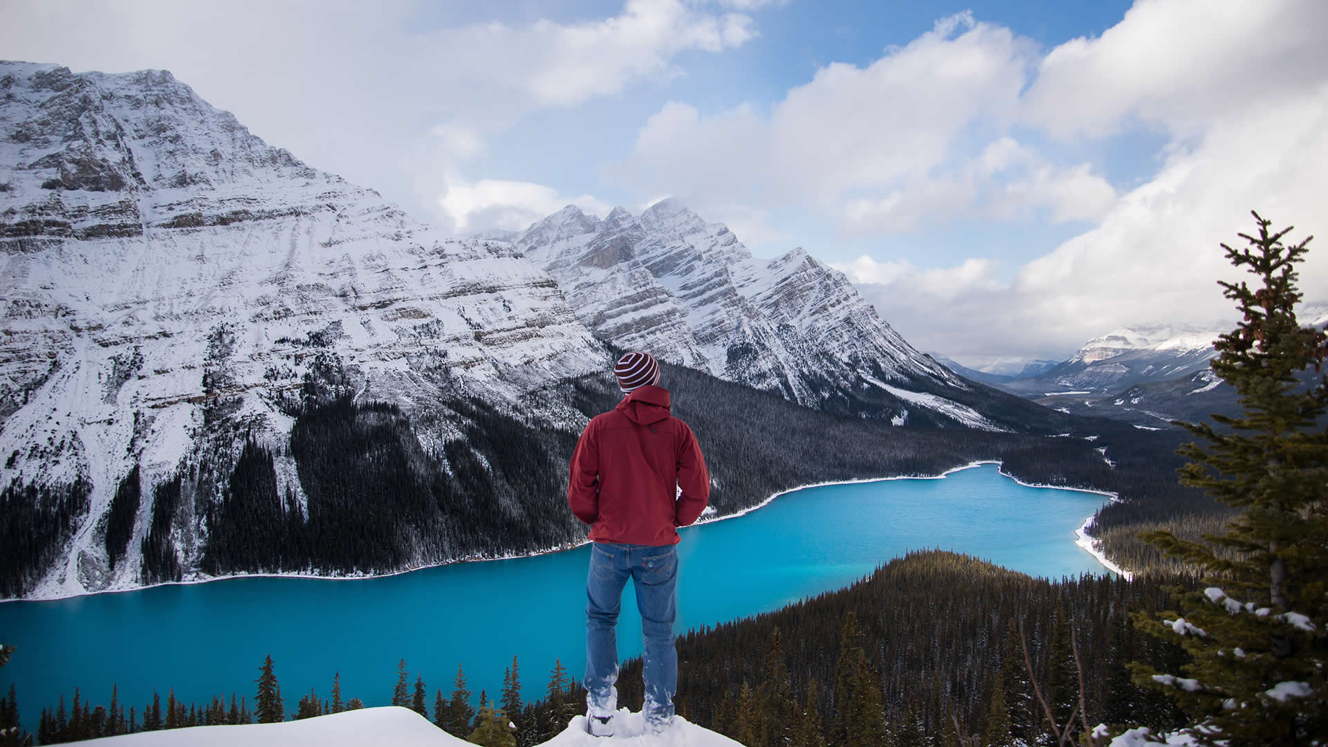 A man stands with the world at his feet, surrounded by snowy mountains and a sparkling blue lake