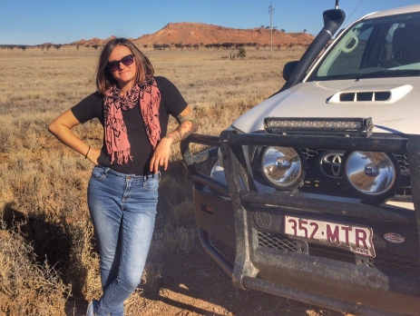 Alysha leaning on the bonnet of a ute with a rugged outback scene of rocks and dessert in the background 