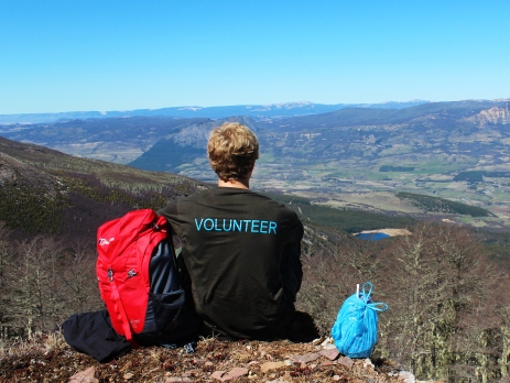 Volunteer with his back to the camera wearing an Oyster t-shirt looks out over the mountains of Chile. There is a backpack to his left and bag to his right as he sits