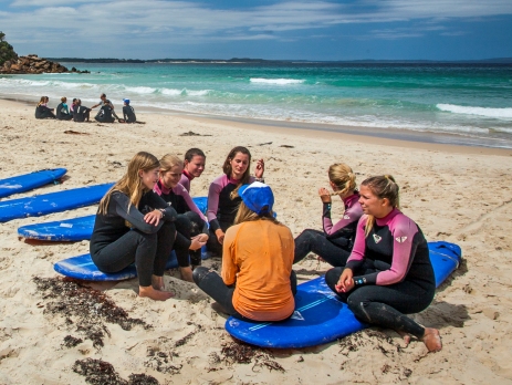 Group of surf students sitting on boards on the beach learning from an instructor with the ocean as the backdrop