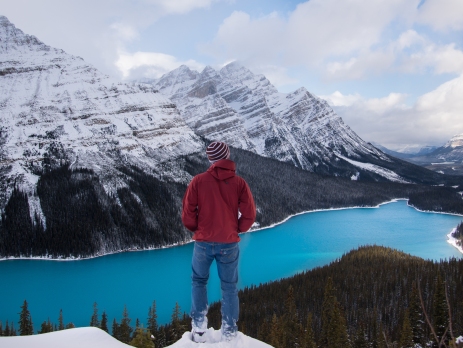 Travellers looks down towards a crystal blue river from a snowy mountain peak. Traveller has their back to the camera